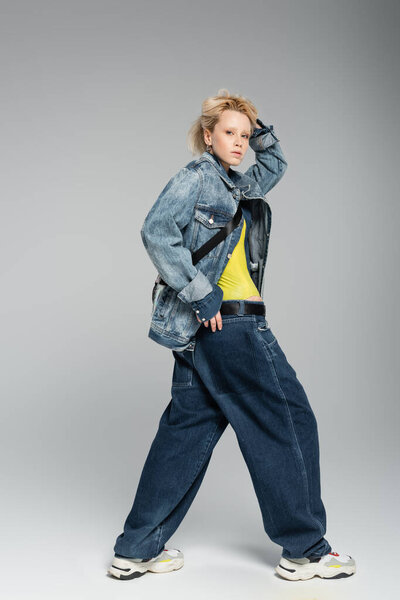 full length of young blonde model in stylish denim outfit and trendy sneakers posing on grey 
