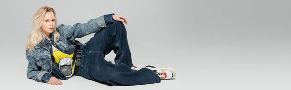 full length of young blonde woman in blue denim outfit and trendy sneakers posing on grey background, banner