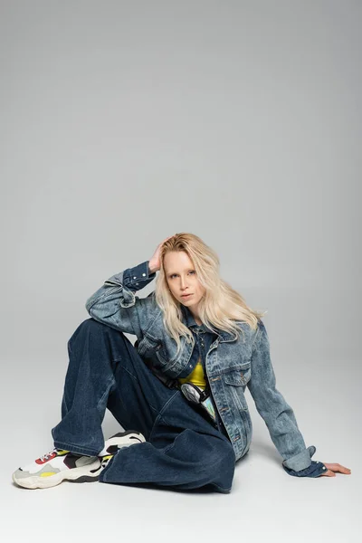 full length of young blonde woman in blue denim outfit and trendy sneakers sitting on grey