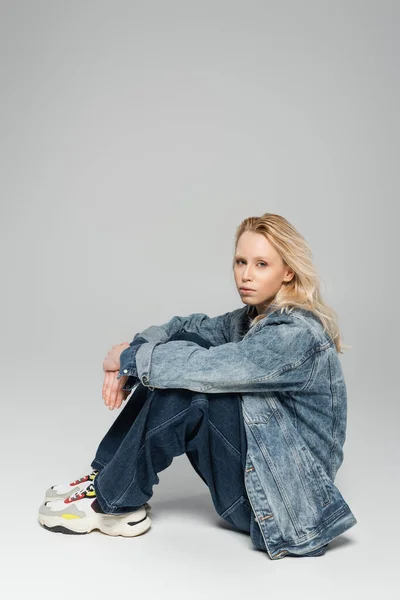full length of blonde albino woman in total denim outfit and trendy sneakers sitting on grey