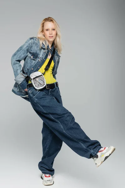 full length of young blonde woman in blue denim outfit and trendy sneakers standing on grey