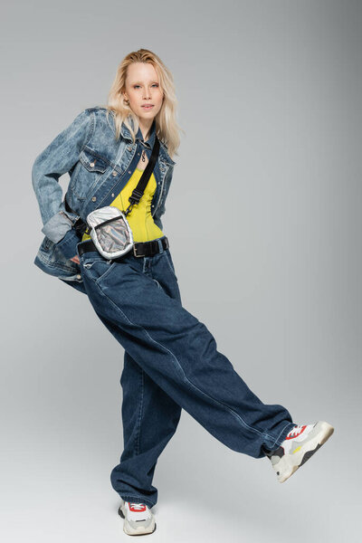 full length of young blonde woman in blue denim outfit and trendy sneakers standing on grey 