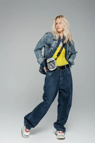 full length of blonde woman in blue denim outfit and trendy sneakers standing on grey
