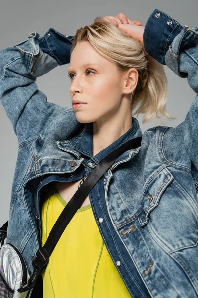 blonde woman in stylish denim jacket posing with hands on head isolated on grey