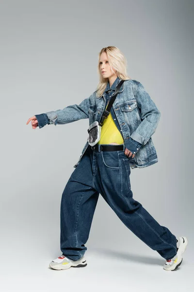 full length of young blonde woman in blue denim outfit and trendy sneakers posing with outstretched hand on grey