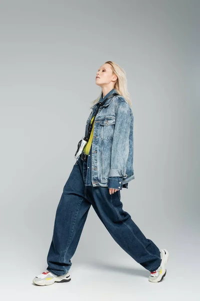full length of young blonde woman in blue denim outfit and trendy sneakers walking on grey