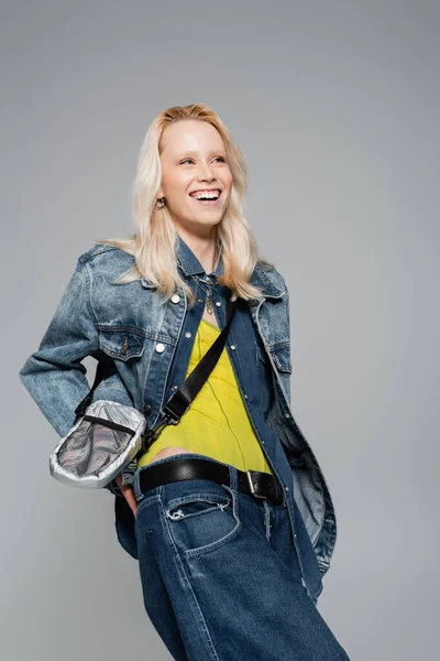 happy woman in denim outfit and belt bag smiling isolated on grey
