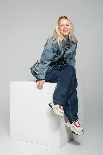full length of cheerful blonde woman in blue denim outfit and trendy sneakers sitting on white cube on grey