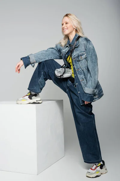full length of happy blonde woman in blue denim outfit and trendy sneakers stepping on white cube on grey