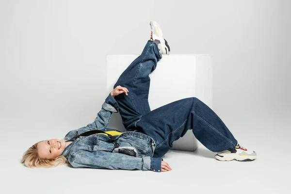 full length of happy blonde woman in blue denim outfit and trendy sneakers lying near white cube on grey