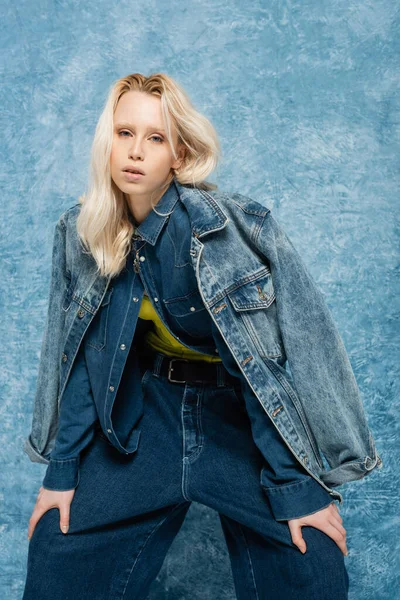 blonde woman in denim jacket posing while looking at camera near blue textured background