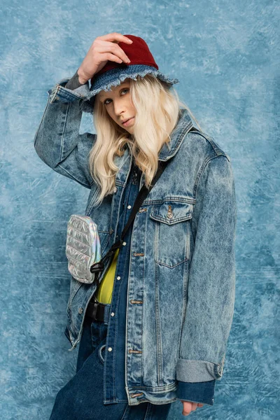 blonde woman in denim jacket and panama hat looking at camera near blue textured background