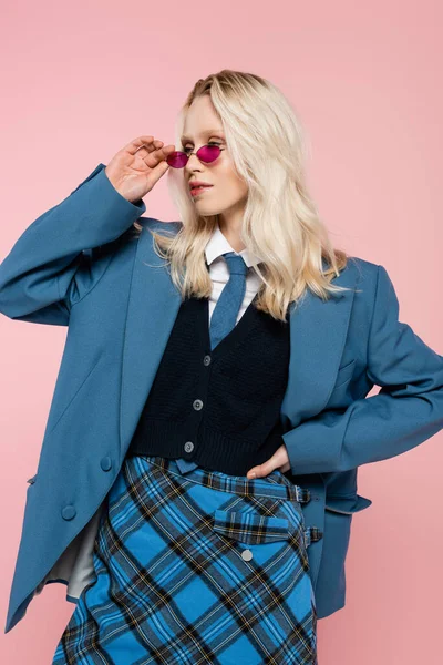 Stock image blonde woman in blue blazer with tie and sunglasses posing with hand on hip isolated on pink 