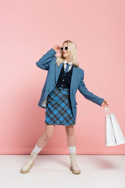 Stock image full length of blonde woman in blue outfit adjusting trendy sunglasses and holding shopping bags on pink 