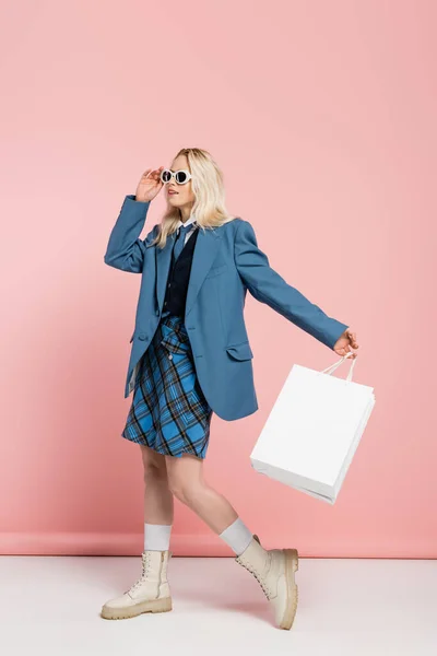 full length of blonde woman in blue blazer adjusting trendy sunglasses and holding shopping bags on pink