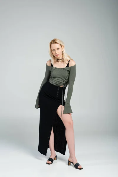 full length of blonde woman in long sleeve and sexy black skirt posing on grey