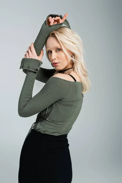 blonde woman in long sleeve shirt and black skirt looking at camera while posing isolated on grey