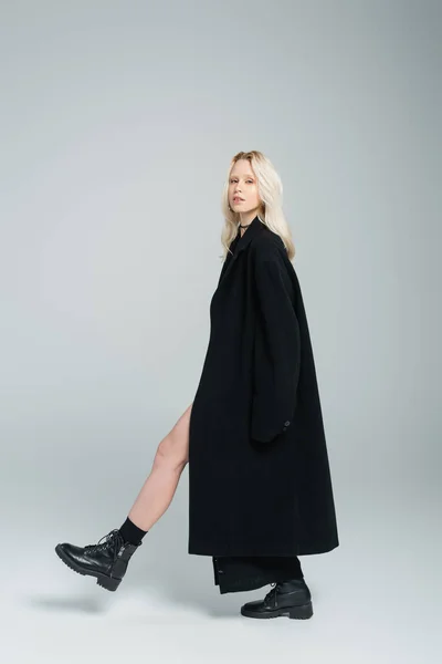 full length of blonde young woman in black coat walking and looking at camera on grey