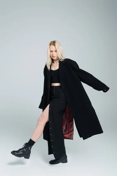full length of blonde woman in black coat and skirt looking at camera while posing on grey