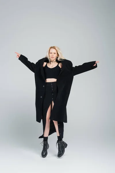 full length of blonde and young woman in stylish black outfit levitating on grey