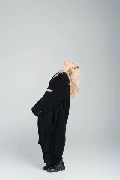 full length of blonde young woman in stylish black outfit standing with raised head on grey