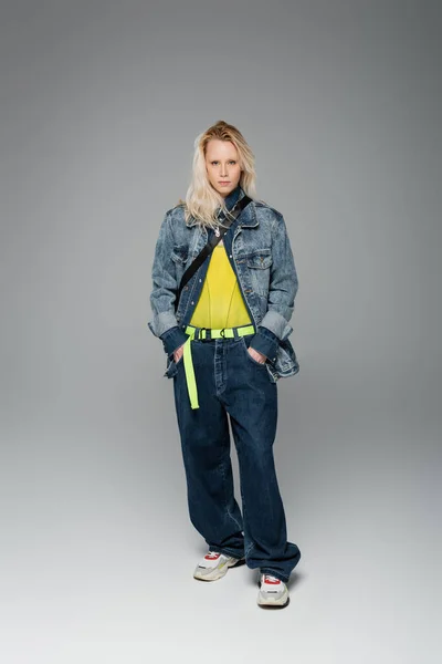 full length of blonde albino woman in blue denim outfit posing with hands in pockets on grey