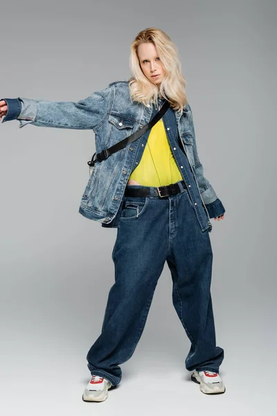 full length of blonde albino woman in blue denim outfit posing with outstretched hand on grey