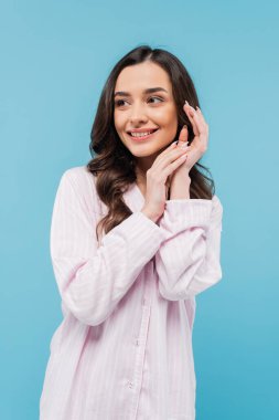 happy young woman with wavy brunette hair standing in pajamas isolated on blue 
