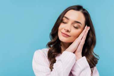 sleepy young woman with closed eyes folding hands while standing in pajamas isolated on blue 