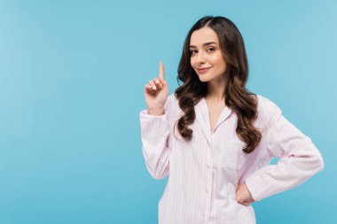 brunette young woman in sleepwear showing idea sign isolated on blue  clipart