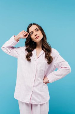 pensive young woman in sleepwear looking away and standing with hand on hip isolated on blue 