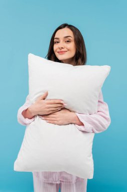 pleased young woman in sleepwear holding white pillow isolated on blue 