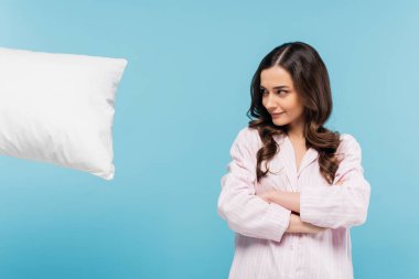 brunette young woman in sleepwear standing with crossed arms and looking at levitating pillow isolated on blue 