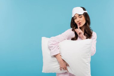 young woman in nightwear and sleeping mask holding white pillow while showing hush sign isolated on blue 