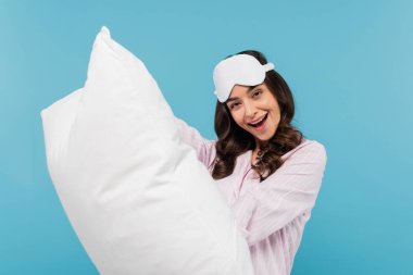 excited woman in nightwear and sleeping mask holding white pillow isolated on blue 