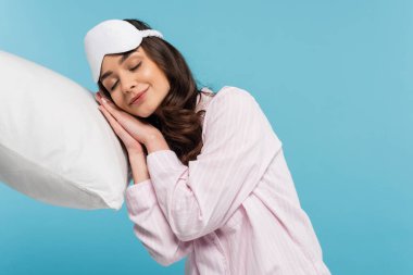 pleased young woman in pajamas and night mask sleeping on white pillow isolated on blue 