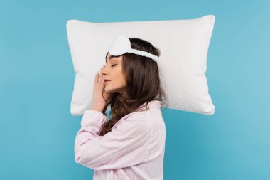 side view of brunette woman in pajamas and night mask sleeping on white pillow isolated on blue 