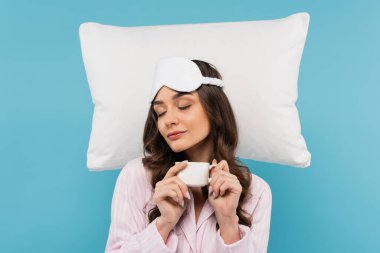 Pijamalı ve gece maskeli uykulu bir kadın, mavi üzerine izole edilmiş beyaz uçan yastığın yanında bir fincan kahve tutuyor.