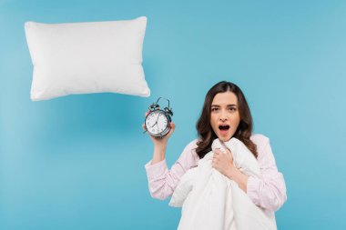 shocked young woman in pajamas holding duvet and vintage alarm clock near flying pillow on blue  clipart