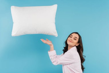 happy woman in pajamas pointing at white flying pillow in air on blue background 