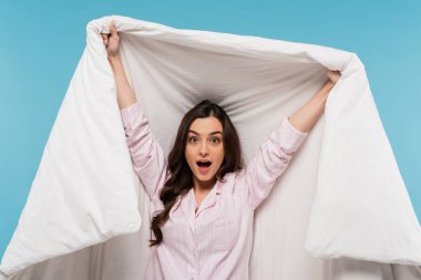 amazed woman in pajamas holding warm duvet and looking at camera isolated on blue  clipart