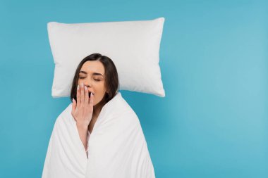 tired woman covered in white duvet standing near flying white pillow and yawning on blue background  clipart