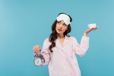 brunette young woman in night mask and sleepwear holding alarm clock and cup isolated on blue  clipart