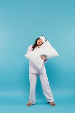 full length of sleepy young woman in night mask and pajamas holding pillow on blue 