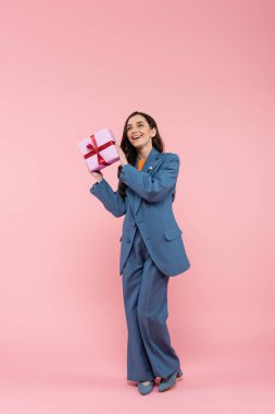 full length of happy woman in blue pantsuit holding wrapped present with ribbon on pink 