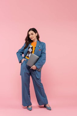 full length of young brunette freelancer in blue pantsuit holding laptop while posing with hand in pocket on pink 