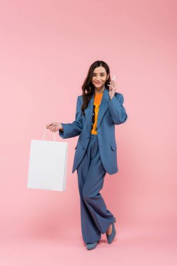 full length of smiling woman in blue pantsuit talking on smartphone and holding shopping bag on pink 