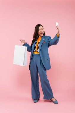 full length of excited woman in blue pantsuit holding shopping bag and taking selfie on pink  clipart