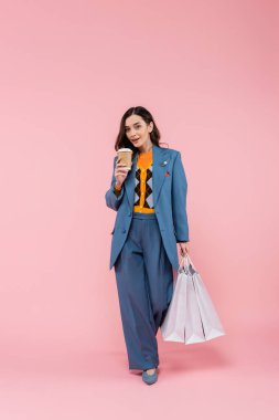 full length of brunette woman in blue suit holding shopping bags and coffee to go on pink 