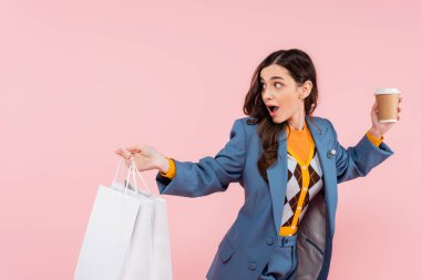 shocked young woman in blue blazer holding shopping bags and coffee to go isolated on pink 